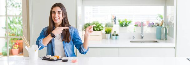 Laaja kulma kuva kaunis nuori nainen syö toimitus sushi hymyillen ja katsomalla kameraa osoittaa kahdella kädellä ja sormella sivulle
. - Valokuva, kuva