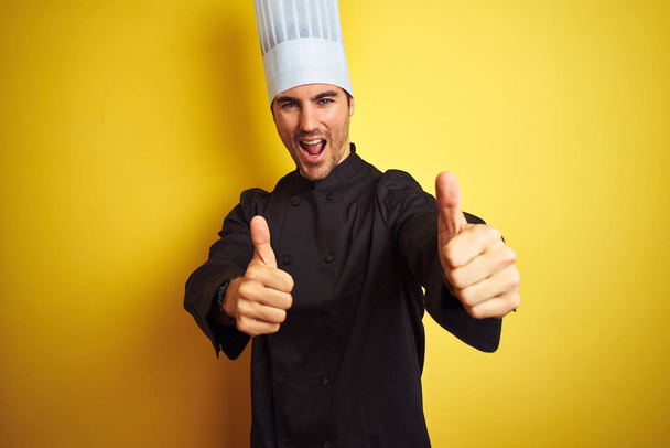 Young chef man wearing uniform and hat standing over isolated yellow background approving doing positive gesture with hand, thumbs up smiling and happy for success. Winner gesture. - Фото, зображення