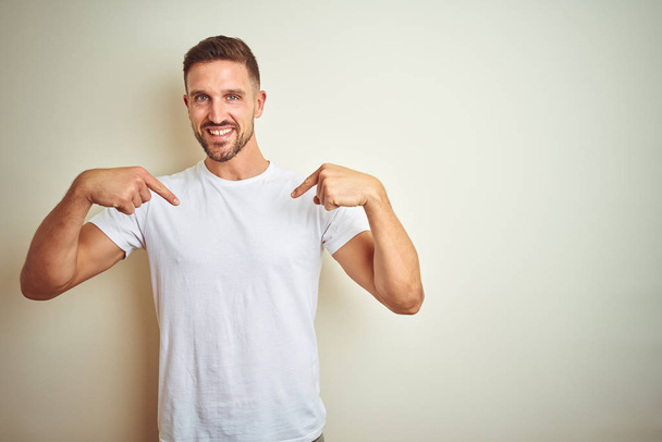 Young handsome man wearing casual white t-shirt over isolated background looking confident with smile on face, pointing oneself with fingers proud and happy. - Photo, Image