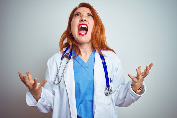 Nuori punapää lääkäri nainen käyttää stetoskooppi yli valkoinen eristetty tausta hullu ja hullu huutaa ja huutaa aggressiivinen ilme ja kädet ylhäällä. Turhautumisen käsite
. - Valokuva, kuva