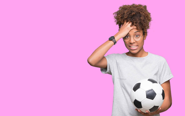 Молодая африканская американка, держащая футбольный мяч на изолированном фоне, напряженная рукой на голове, потрясенная стыдом и удивлением, сердитая и разочарованная. Страх и разочарование по ошибке
. - Фото, изображение