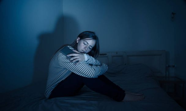 Young teenager girl suffering mobile cell phone addiction feeling lonely and depressed having insomnia needing to be connected sitting on bed late at night. In - Фото, изображение