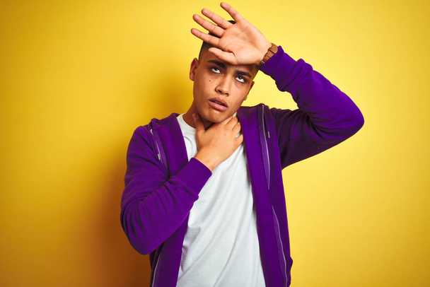 junger brasilianischer Mann in lila Sweatshirt, der vor isoliertem gelben Hintergrund steht und die Stirn vor Krankheit und Fieber, Grippe und Erkältung, Viruserkrankung berührt - Foto, Bild
