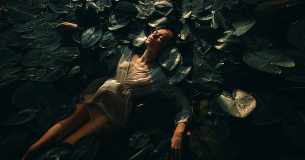 Mujer joven yaciendo en el agua entre las hojas y flores de y
 - Foto, Imagen