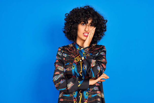 Young arab woman with curly hair wearing colorful shirt over isolated blue background thinking looking tired and bored with depression problems with crossed arms. - Photo, Image