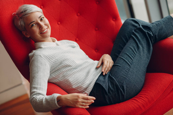 Porträt einer stilvoll lächelnden jungen Frau im weißen Pullover, die im roten Stuhl sitzt - Foto, Bild