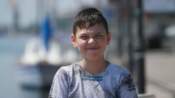 Happy boy standing and smiling cheerily on Dnipro quay in summer in slo-mo            Cheerful view of a smiling brunette boy with short haircut in colorful T-shirt standing and enjoying his life on Dnipro river embankment in summer in slow motion - Footage, Video