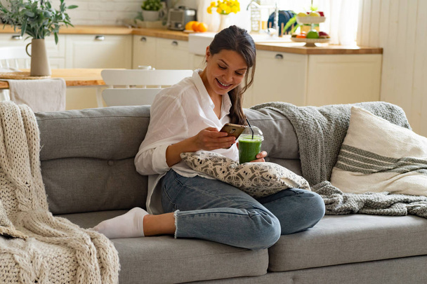 happy woman smiling while using smartphone and sitting on couch with cup of green smoothie at home - Zdjęcie, obraz
