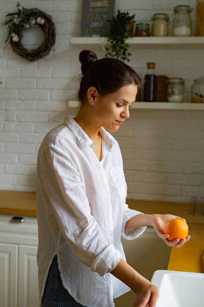Young woman holding fresh orange while standing at kitchen  - Foto, imagen