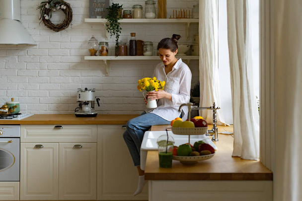 Young woman holding vase with bright yellow flower bouquet while sitting on table at kitchen - Foto, Bild