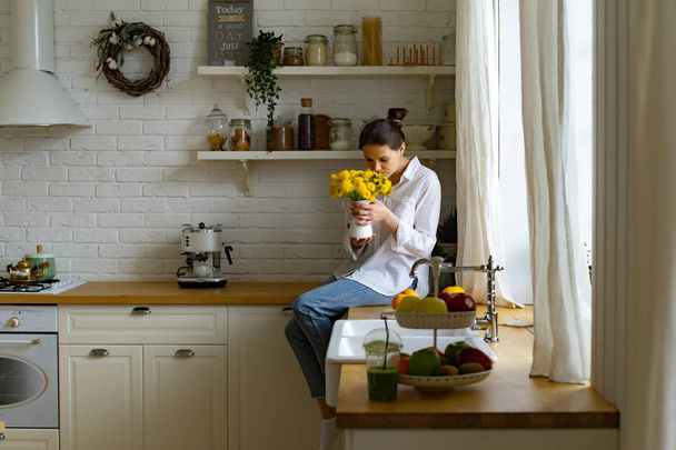 Jeune femme tenant vase avec bouquet de fleurs jaune vif tout en étant assis sur la table à la cuisine
 - Photo, image