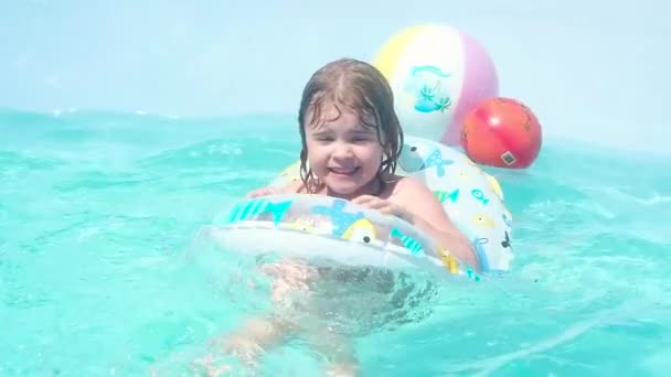 malá krásná Kavkazská dívka s vlhkýma tmavými vlasy plavá na nafukatelný kruh dětí v bazénu. Aktivně vytahuješ nohy pod vodu, zamhouřil se před šploucháním a vesele se zasmál. - Záběry, video
