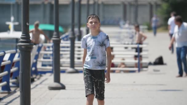 Jolly boy walking in the Dnipro river quay on a sunny day in summer in slo-mo           Cheery view of happy brunette boy with short haircut in T-shirt walking with inspiration and smiling in the Dnipro quay with yachts and boats in summer in slo-mo - Video