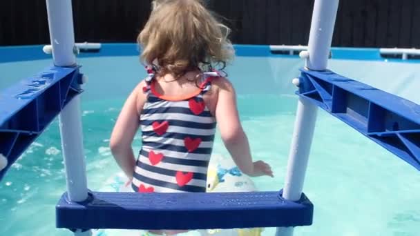 kleines Mädchen im Badeanzug mit roten Herzen springt mit einem aufblasbaren Kreis von der Pooltreppe und beginnt aktiv zu schwimmen und hat Spaß dabei, sich die Zunge herauszulachen. Schwimmen im heimischen Teich an heißen Sommertagen. - Filmmaterial, Video