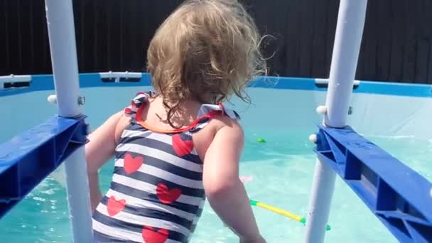 little girl with light wet hair in swimsuit with red hearts, looks at camera and jumps with an inflatable circle from pool stairs and begins to swim actively. Swimming in  home pond on hot summer day. - Footage, Video