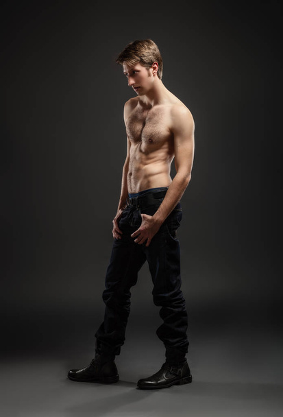 Ολόσωμο πορτραίτο ενός σέξι νεαρού άντρα με τζιν και ημίγυμνος που ποζάρει στο στούντιο. - Φωτογραφία, εικόνα