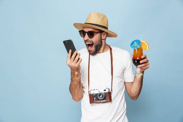 Excited young man tourist posing with camera using mobile phone drinking cocktail isolated over blue wall background. - 写真・画像