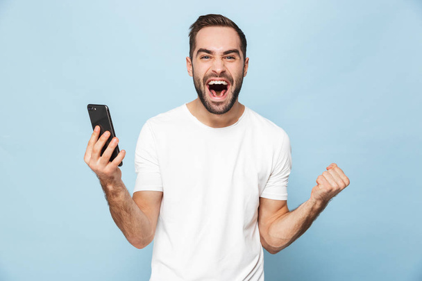 Handsome excited young man using mobile phone isolated over blue wall background. - Фото, зображення
