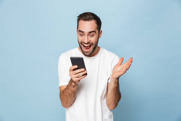 Handsome excited young man using mobile phone isolated over blue wall background. - Photo, image