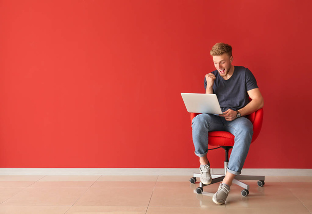 Счастливый молодой человек с ноутбуком сидит на стуле рядом с цветной стеной
 - Фото, изображение