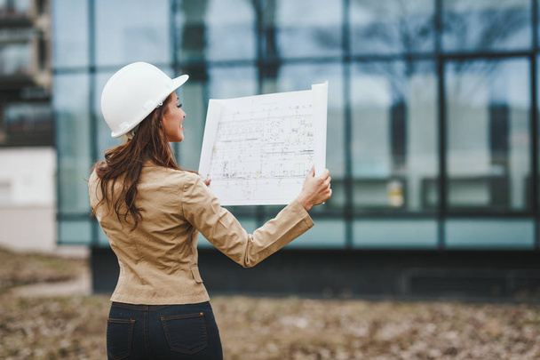 Portrait of young engineer woman in white helmet holding blueprints at construction site - Fotó, kép