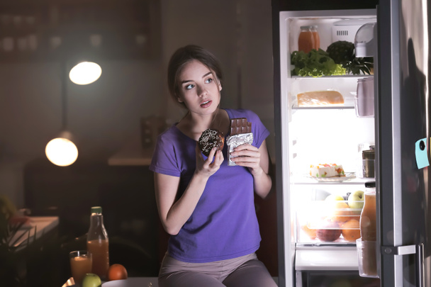 Настороженная молодая женщина ест нездоровую пищу возле холодильника на кухне ночью
 - Фото, изображение