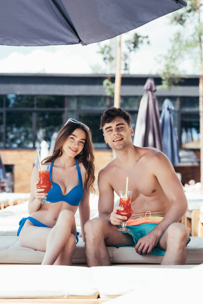 cheerful young couple sitting on deck chair with glasses of refreshing drink and looking at camera - Photo, Image