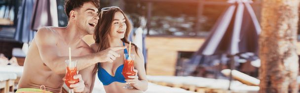panoramic shot of cheerful young man pointing with finger while sitting near pretty girlfriend with glasses of refreshing drink - Photo, Image