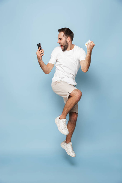 Photo of positive caucasian man in casual white t-shirt rejoicing while holding credit card and smartphone - Fotoğraf, Görsel