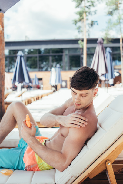 handsome young man applying sunscreen lotion while sitting on chaise lounge - Photo, image