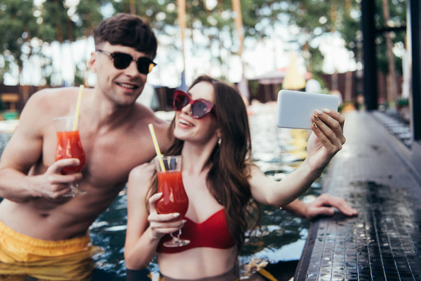alegre joven mujer tomando selfie con novio mientras se relaja en la piscina juntos
 - Foto, Imagen
