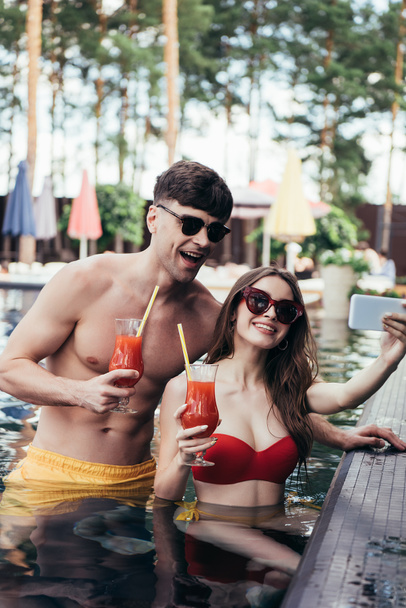 cheerful couple holding glasses of refreshing drink and taking selfie while resting in swimming pool - Photo, Image