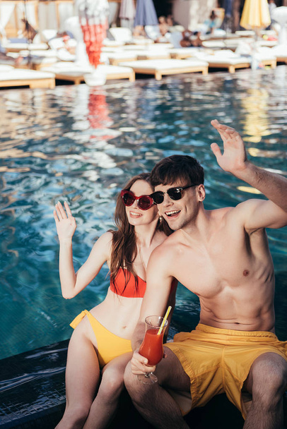 cheerful young couple looking away and waving hands while sitting at poolside - Foto, imagen