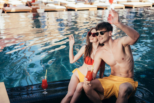 happy young couple looking away and waving hands while sitting at poolside - Фото, изображение