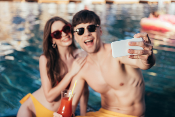 enfoque selectivo de alegre pareja joven tomando selfie mientras está sentado en la piscina
 - Foto, Imagen