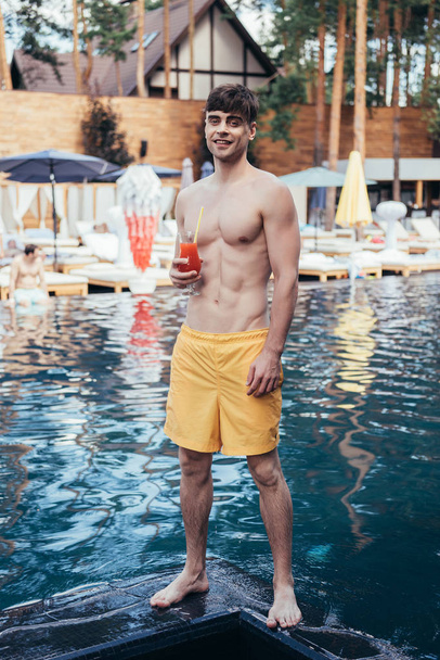 serinletici içecek cam ile yüzme havuzu yakınında dururken kameraya gülümseyen yakışıklı genç adam - Fotoğraf, Görsel