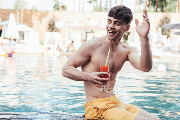 cheerful young man looking away and waving hand while relaxing at poolside with glass of refreshing beverage - Photo, Image