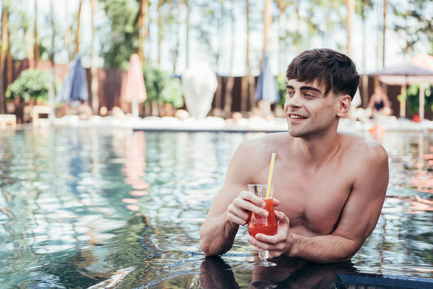 smiling young man looking away while relaxing in swimming pool with glass of refreshing drink - Foto, afbeelding