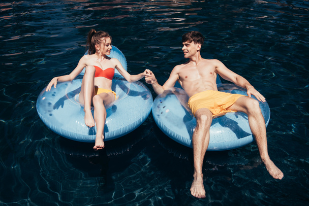 Szczęśliwa Młoda para trzyma ręce podczas relaksu na ringach pływackich w basenie - Zdjęcie, obraz