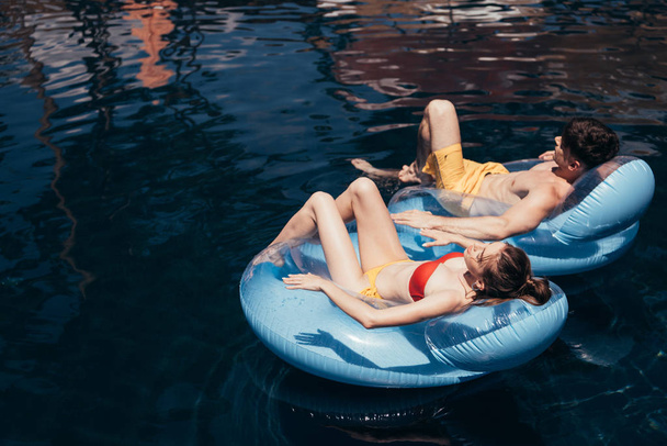 young couple sunbathing while floating on swim rings in swimming pool - Photo, Image