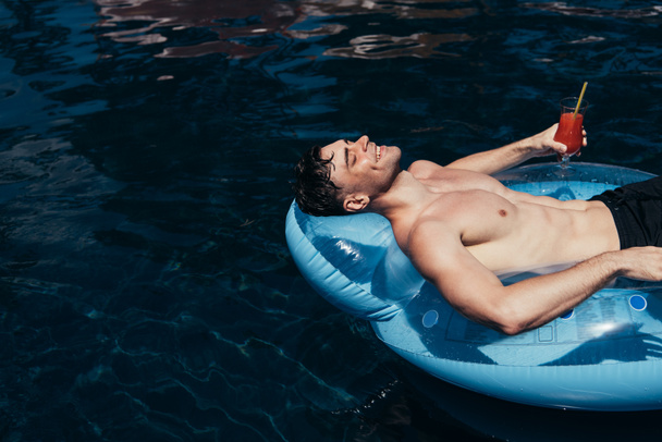 šťastný mladý muž relaxující s plaveckým kroužkem v bazénu se sklenicí osvěžující nápoje - Fotografie, Obrázek