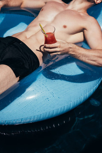 częściowy widok młodego mężczyzny relaksującego się na pierścieniu pływającym w basenie ze szklanką orzeźwiającego napoju - Zdjęcie, obraz