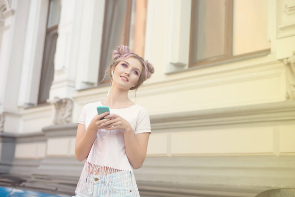 Portrait of a young stylish hipster woman walking down the street dressed in a trendy outfit, a pink hairstyle bun traveling around holding a mobile phone. - Valokuva, kuva