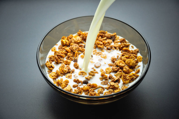 A plate bowl of cereal oatmeal and milk. Milk is pouring in a plate on a dark background. Healthy breakfast. Proper nutrition. - Photo, Image