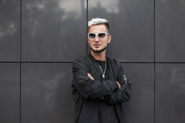 Modern young stylish man model in fashionable black jacket in trendy sunglasses in t-shirt with a hairstyle posing near a modern gray wall in the city. Attractive guy blond hipster. Street style. - Photo, Image