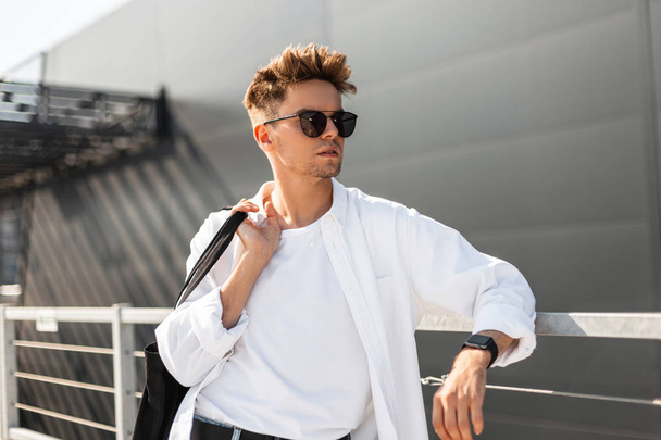 Cool joven hipster hombre en una camiseta blanca con una camisa elegante en gafas de sol de moda está de pie cerca de una valla de metal en una calle de la ciudad. Moderno chico modelo disfruta relajarse en un día soleado
. - Foto, imagen
