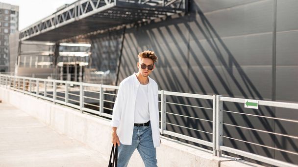 Modern young man hipster with stylish hairstyle in fashionable summer clothes with a black cloth bag in vintage sunglasses walks around the city near a gray building on a sunny day. European guy.  - Zdjęcie, obraz