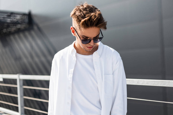 Przystojny nowoczesny młody człowiek w modne białe ubrania w stylowych czarnych okularów przeciwsłonecznych z modną fryzurę stoi na jasnym słonecznym dniu w pobliżu szarego budynku w mieście. Amerykański facet model. Styl letni - Zdjęcie, obraz