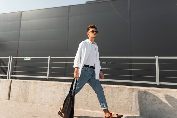 Młody stylowy człowiek Hipster w modne ubrania w okulary przeciwsłoneczne z czarnym tkaniny torba w Vintage sandały spacery jest w mieście w pobliżu szarego budynku. Amerykański facet. Modna letnia Odzież męska. Styl uliczny. - Zdjęcie, obraz