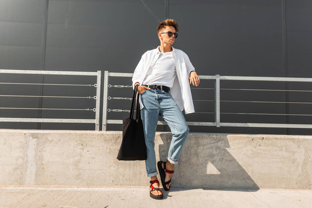 Ein urbaner junger Mann mit Hipster-Frisur in modischer Kleidung und einem Taschenständer neben dem Metallgeländer in der Nähe des grauen Gebäudes. trendiger moderner Kerl. Europäische Jugendmode. - Foto, Bild